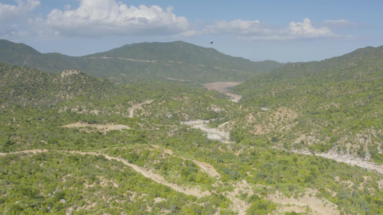 Wide La Palma aerial frame showing layered green hills, a central sandy valley, light dirt roads, distant mountains, scattered clouds, and no visible people.