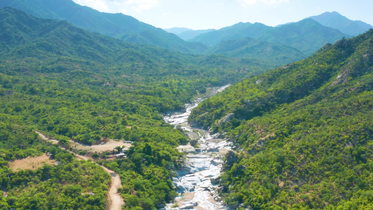 Second aerial frame in La Palma, Baja California Sur, with the white rock channel centered between dense green vegetation, layered mountains beyond, and no visible people.