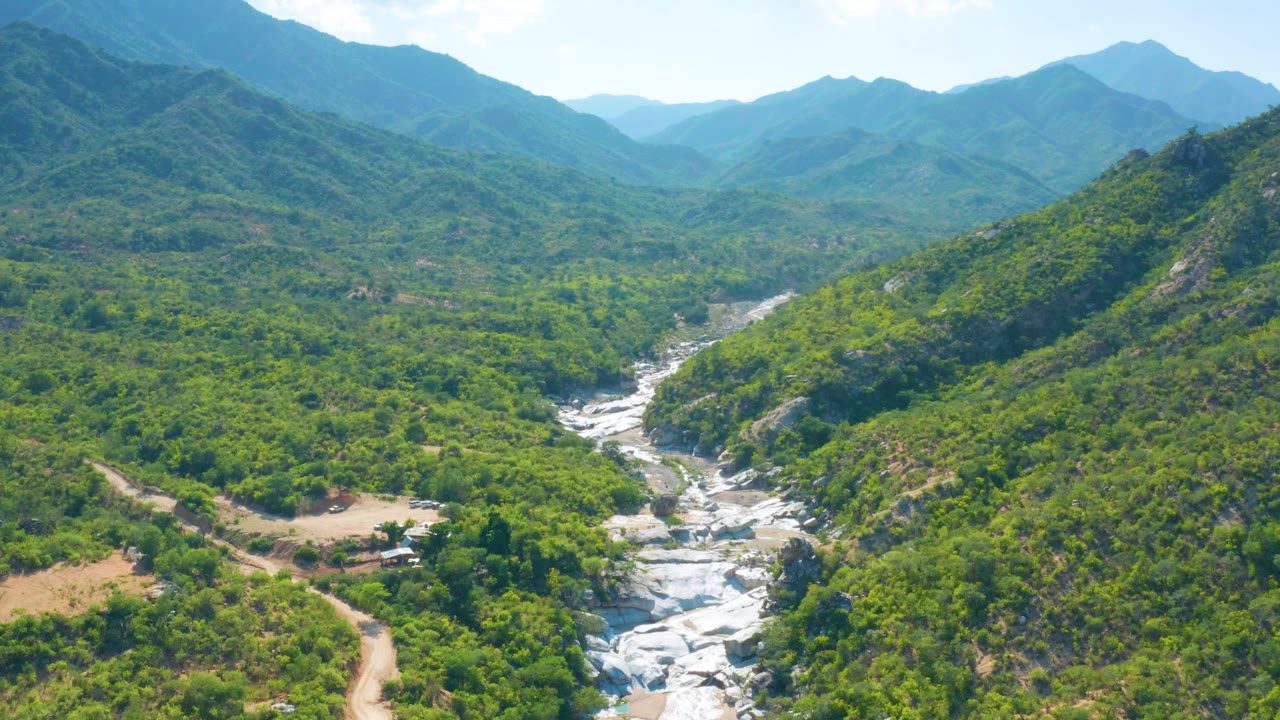 Wide aerial view in La Palma, Baja California Sur, with dense green valley vegetation, pale exposed rocks in the drainage, a ridge rising at right, distant mountains, and no visible people.