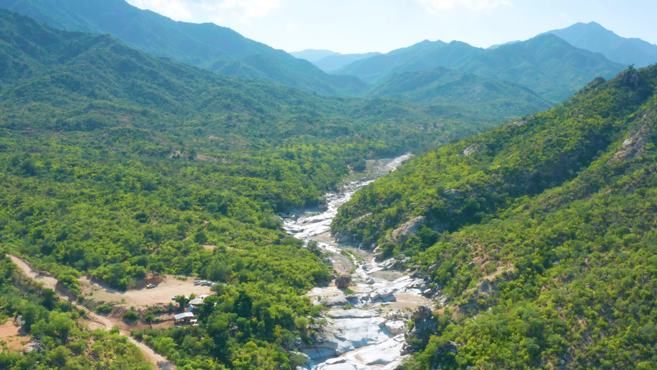 Aerial view in La Palma, Baja California Sur with the pale riverbed slightly more centered between vegetated slopes, distant layered mountains, and no visible people.