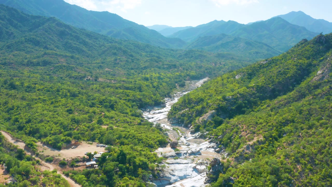Wide aerial frame over La Palma, Baja California Sur showing a rocky waterway in the lower center, dense green vegetation across the valley, hazy blue ridges, and no visible people.