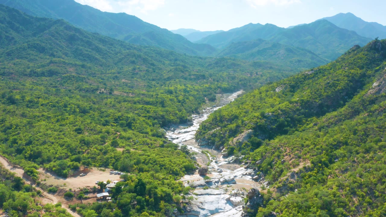 Aerial view in La Palma, Baja California Sur where the river appears closer in the lower frame, bordered by thick green vegetation, sloped hills, distant peaks, and no visible people.