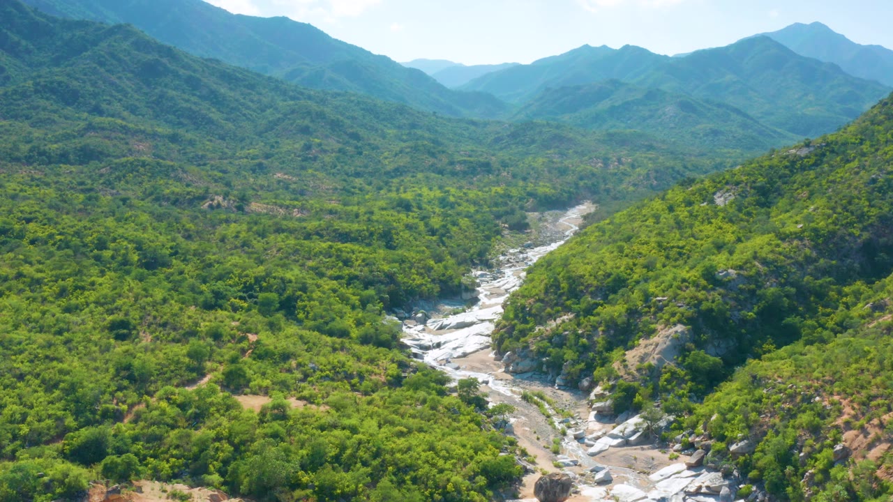 Aerial view in La Palma, Baja California Sur showing exposed light rock in the creek channel, dense green vegetation, distant ridges, and no visible people.
