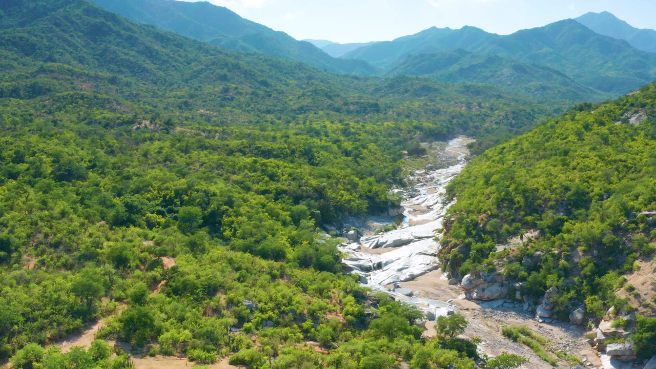Opening aerial frame over La Palma, Baja California Sur, showing green hills around a pale rocky creek bed with layered mountains in the distance and no visible people.