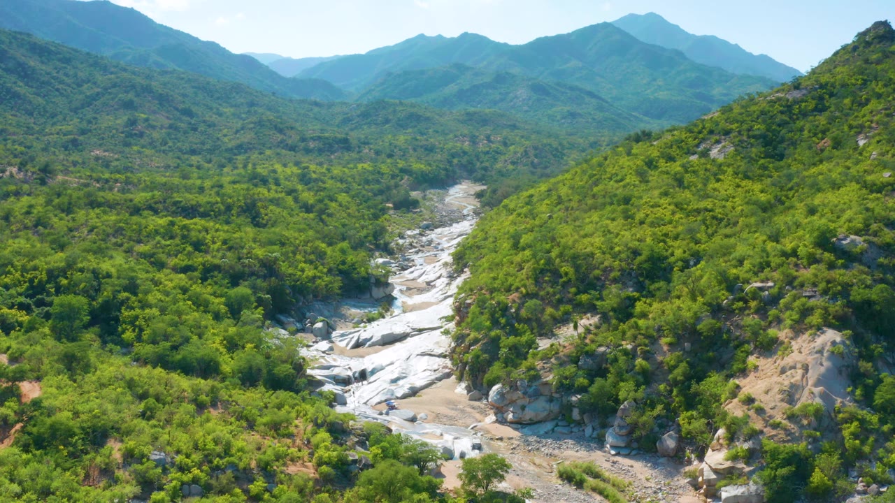 Aerial frame over La Palma, Baja California Sur, Mexico, with white water spread across pale rock below a rounded green ridge and layered mountain background, with no visible people.