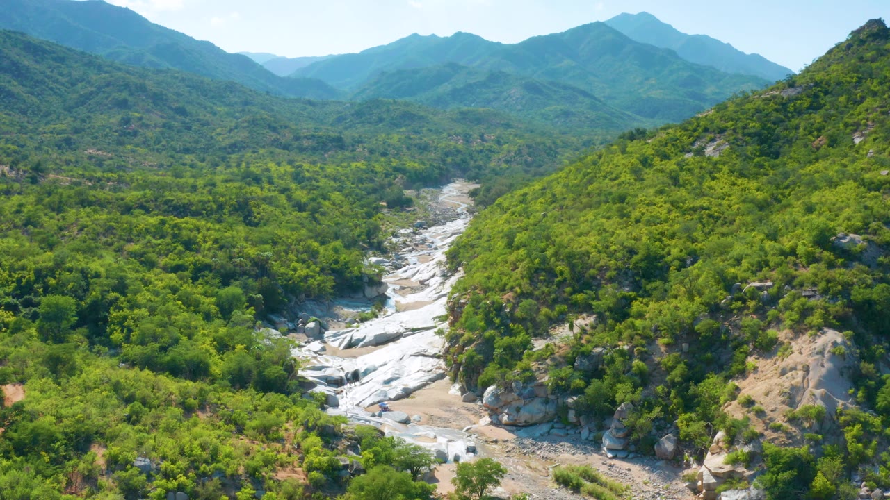 Aerial view in La Palma, Baja California Sur, Mexico, where the rocky riverbed curves through dense green vegetation under a bright sky, with no visible people.