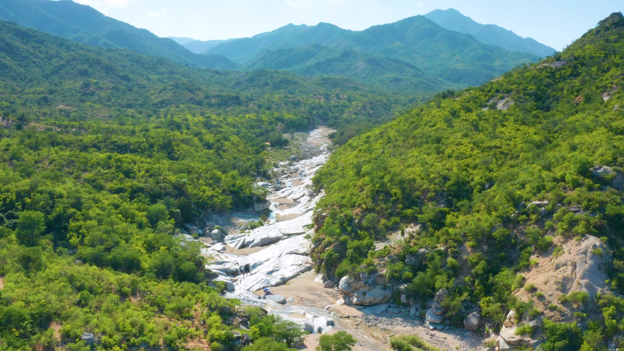 Aerial frame from La Palma, Baja California Sur, Mexico, showing a wide green valley, white river channel, pale rock, and distant mountain layers, with no visible people.