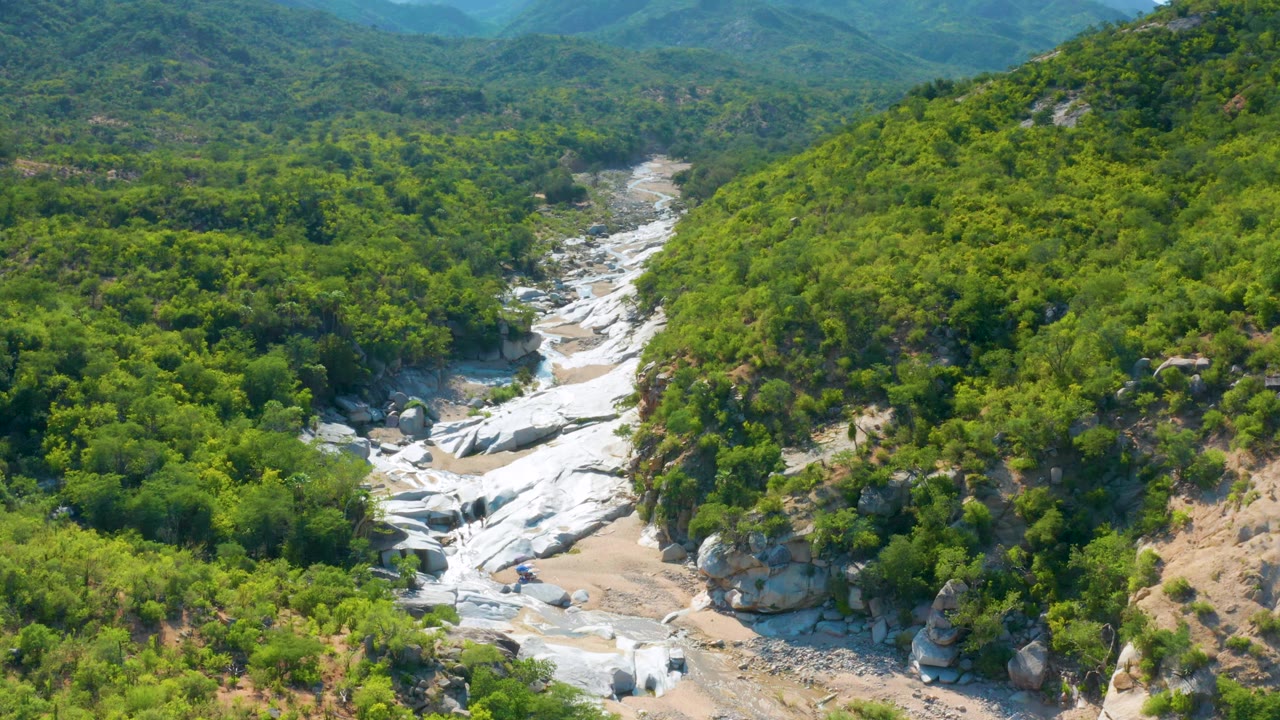 Aerial frame in La Palma, Baja California Sur, with white rock slabs crossing the center of the wash and thick green scrub rising on both sides, no visible people.