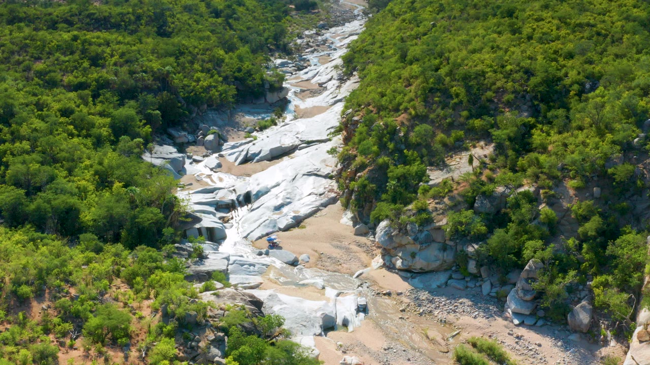 Aerial frame over La Palma, Baja California Sur, showing white rock shelves, sandy wash channels, dense green trees, and tiny people near the lower rocks.