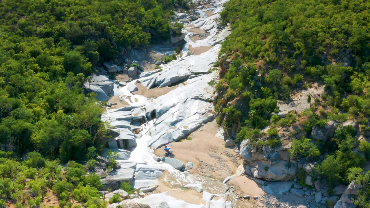 Aerial view in La Palma, Baja California Sur with smooth white rock slabs crossing a sandy dry wash and dense green foliage along both sides, with no visible people.
