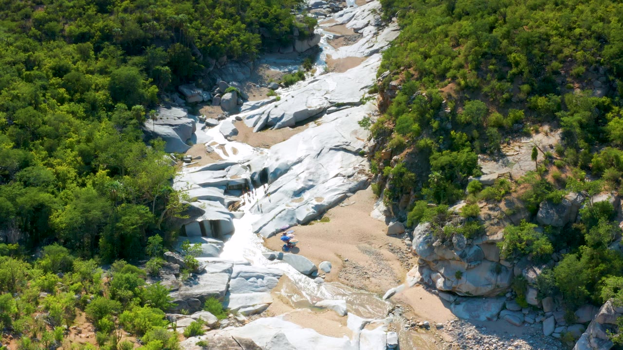 Overhead aerial frame from La Palma, Baja California Sur showing white stone surfaces, tan sand, and thick green vegetation packed around the rock channel, with no visible people.