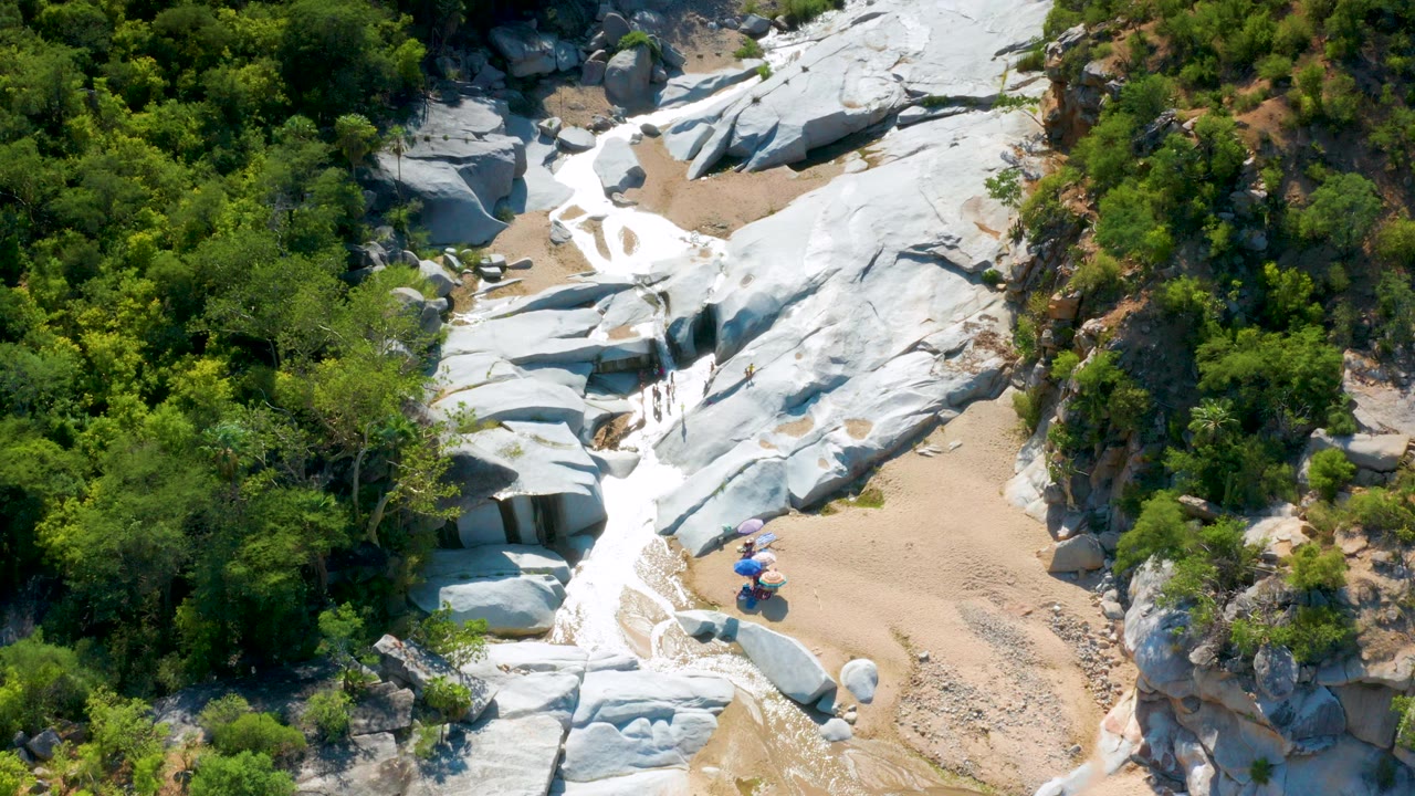 High aerial frame in La Palma, Baja California Sur showing pale rock slabs, a sandy wash, shallow water, dense green brush, and small visible people near blue and purple umbrellas.