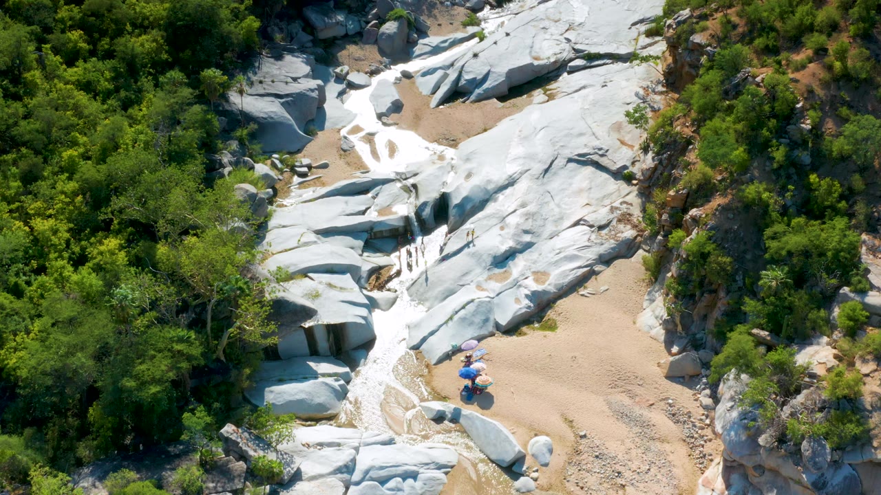 Aerial frame in La Palma, Baja California Sur showing a rocky canyon wash, pale stone surfaces, surrounding green shrubs, and small visible people near umbrellas at the lower center.
