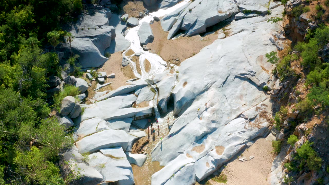 Aerial view in La Palma, Baja California Sur, Mexico, with pale rock slabs, sandy channels, green vegetation, and several small people visible near the lower rock edge.