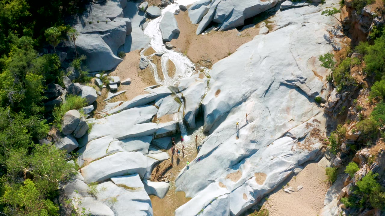 Aerial view in La Palma with large pale rock sheets, a sandy wash at the top, dense green shrubs along the side, and small visitors visible near the lower edge.