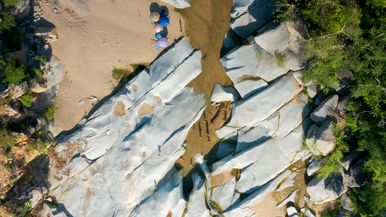 Aerial frame from La Palma, Baja California Sur, Mexico, showing pale rock slabs beside sand, green trees, a narrow water channel, umbrellas near the top edge, and small visible people below.