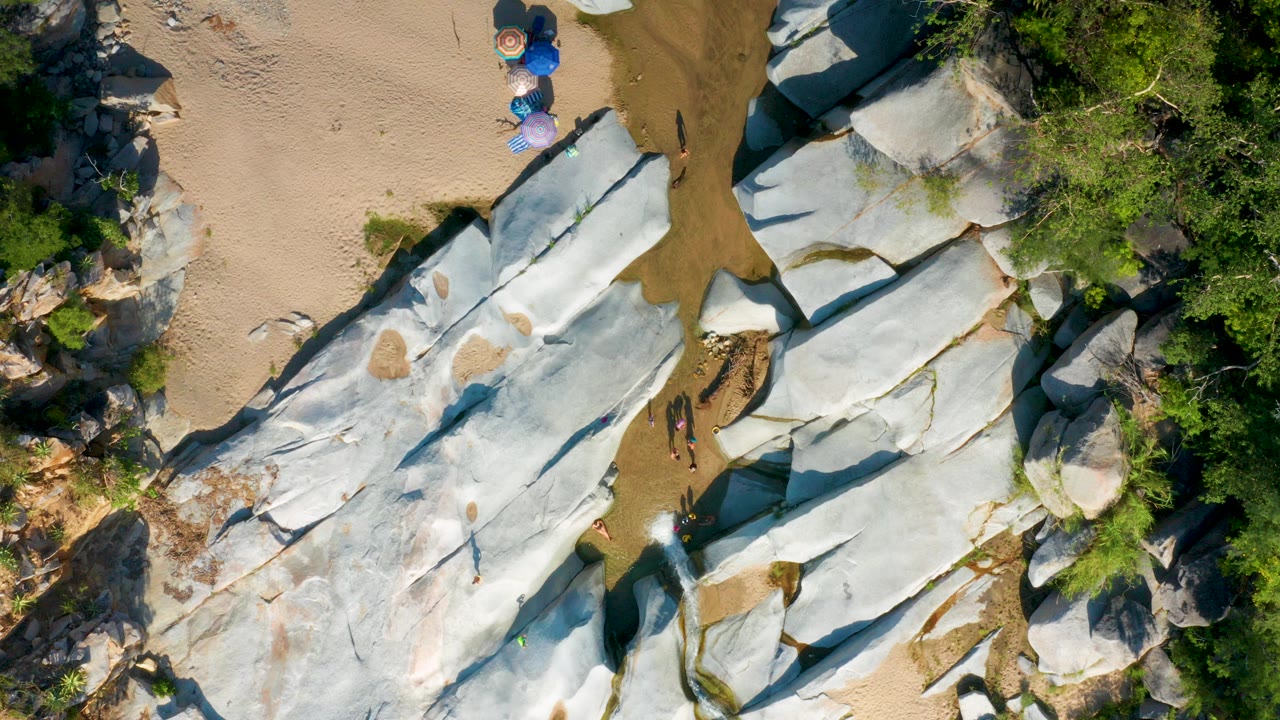 Top-down view in La Palma, Baja California Sur showing pale rock slabs beside tan sand, green plants, beach umbrellas near the upper edge, and tiny distant figures on the rock.
