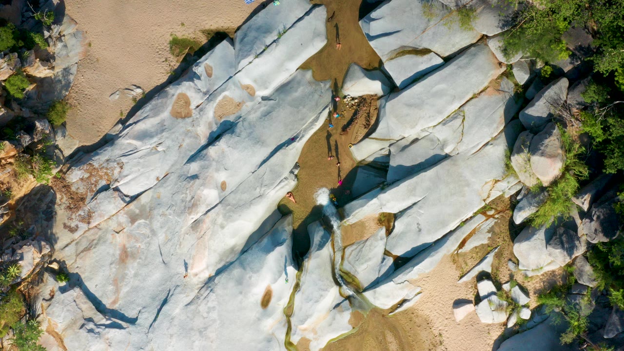 Overhead frame from La Palma, Baja California Sur showing pale grey shoreline rocks, tan sand, sparse green vegetation, and small people visible between the stones.