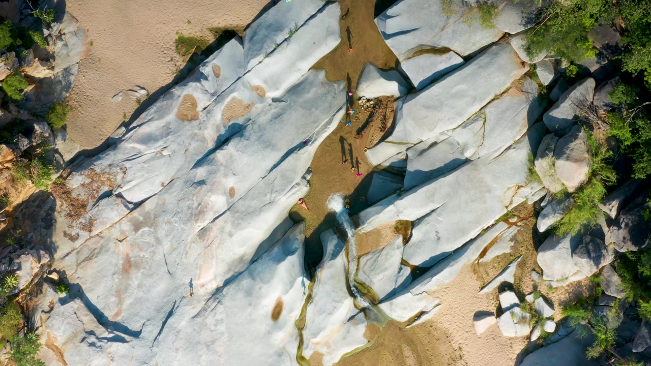 Aerial frame over La Palma shoreline rocks with sandy edges, patches of green vegetation, shallow water in rock seams, and small visible people along the right side.