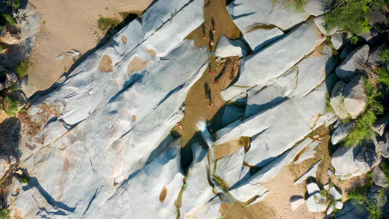 Overhead frame in La Palma, Baja California Sur showing pale rock slabs, tan sand, green shrubs, shallow water channels, and small tourists near the upper center and lower rock edge.