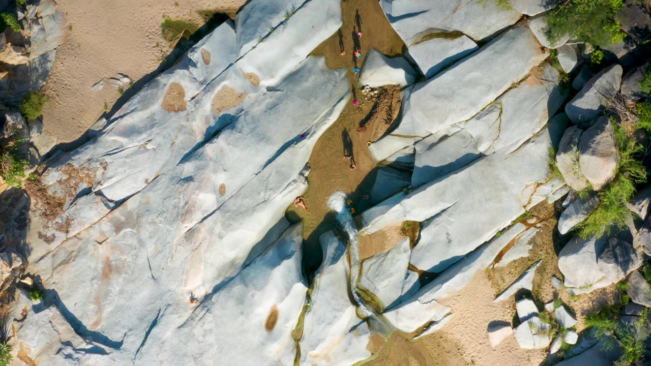 Top-down view in La Palma, Baja California Sur showing a pale stone ridge crossed by dark cracks, tan sand along the upper edge, small tourists near the center, and narrow water pooled in creases.