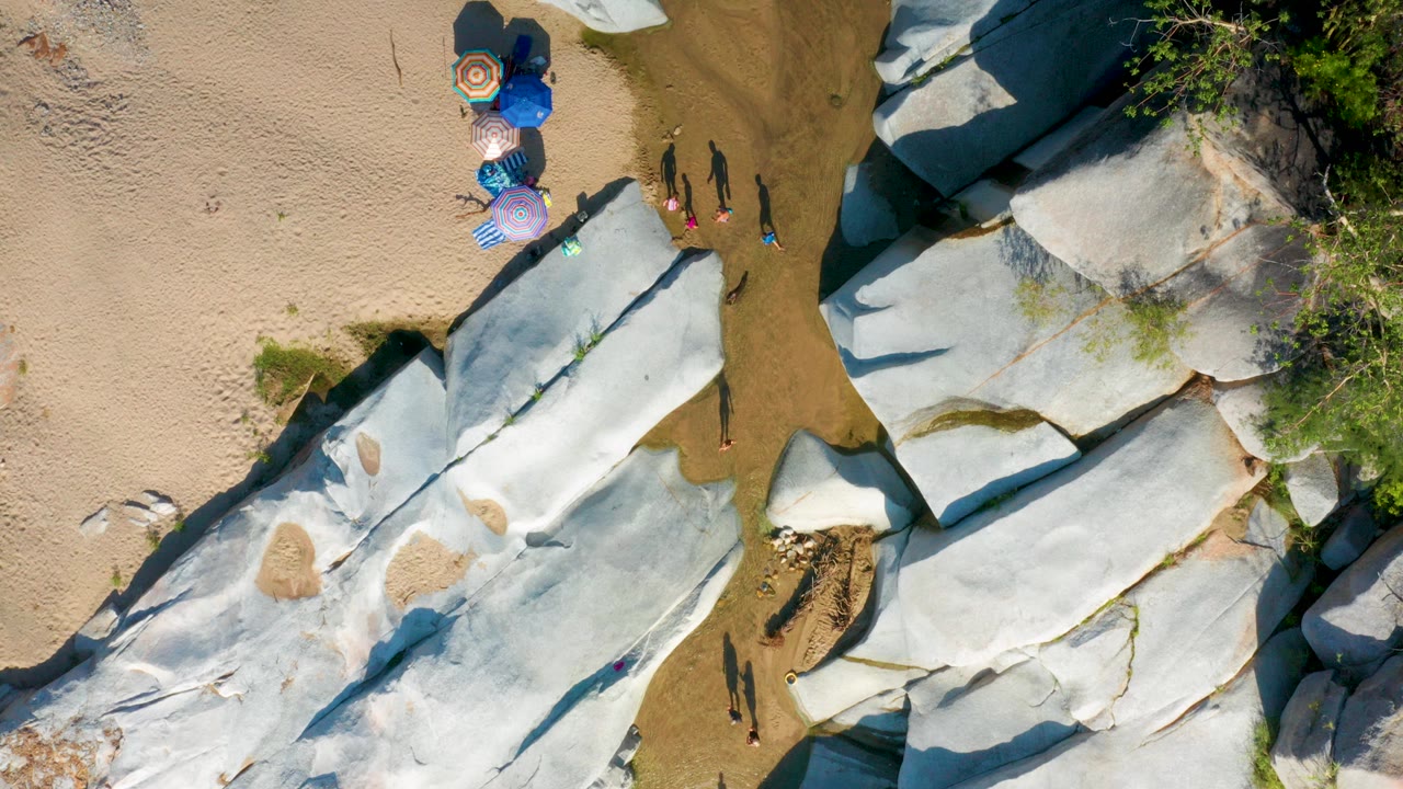 Top-down aerial frame in La Palma, Baja California Sur, showing umbrellas on sand beside pale shoreline rocks, shallow water, green vegetation, and small visible people.