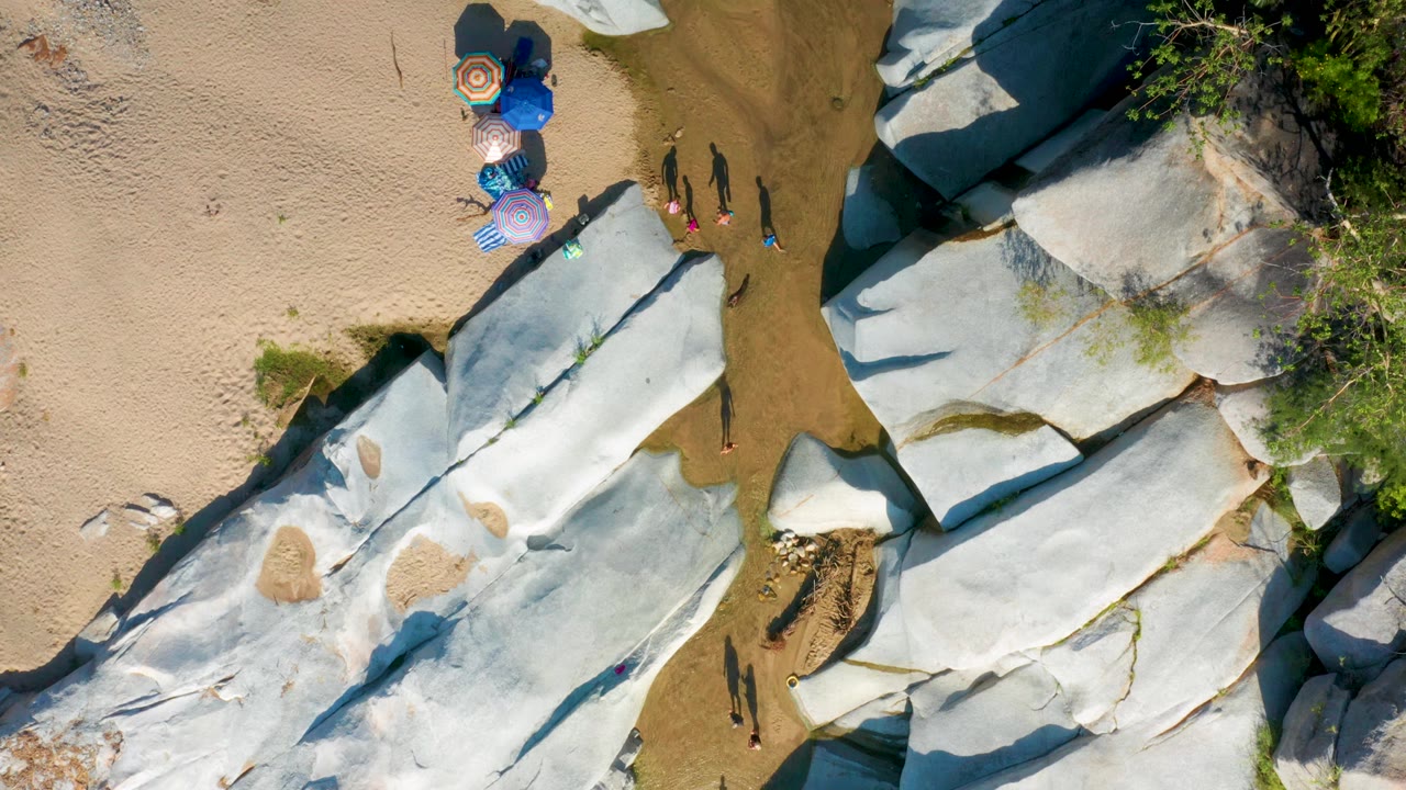 Aerial beach frame with umbrellas near large rocks and people on sand.
