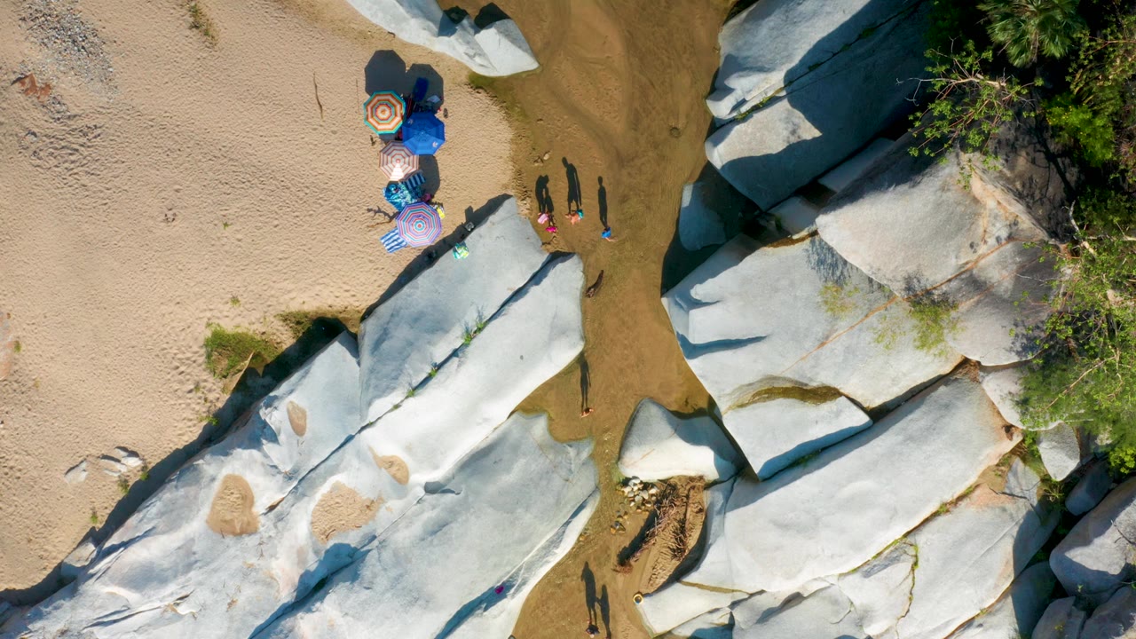 Drone-like overhead frame in La Palma, Baja California Sur, showing the umbrella group near the top of the frame, pale rock slabs below, green vegetation to the side, and small visible people casting shadows.