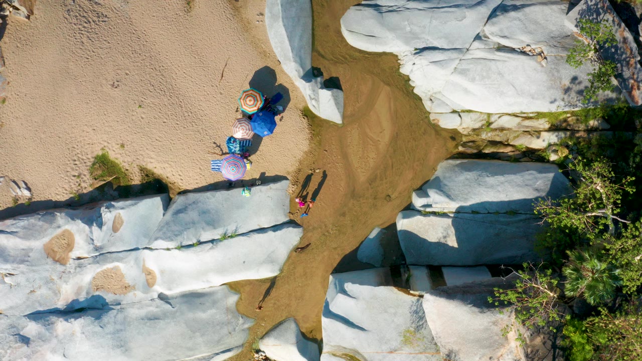 Overhead frame from La Palma, Baja California Sur showing tan sand, pale stone shelves, shallow water, several beach umbrellas, and no recognizable people visible.