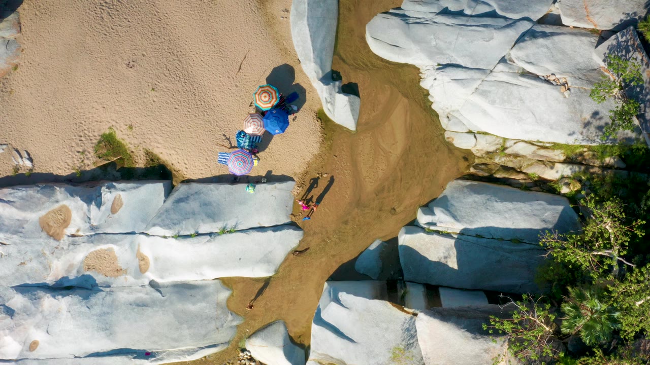 Top-down La Palma frame showing umbrellas on sand above a narrow water-washed area, pale rock slabs on both sides, green vegetation at the lower right, and no recognizable people visible.