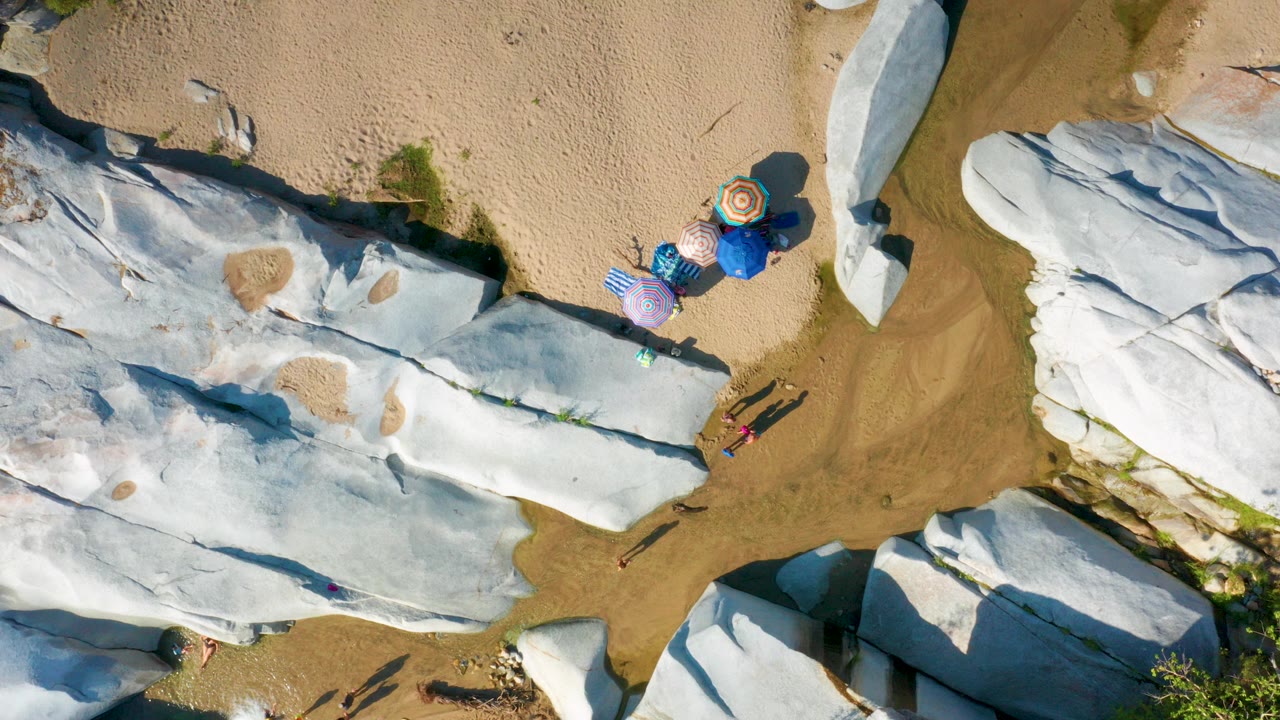 Overhead frame in La Palma, Baja California Sur showing pale rock slabs, tan sand, shallow water, colorful umbrellas, and tiny people near the rock edge.