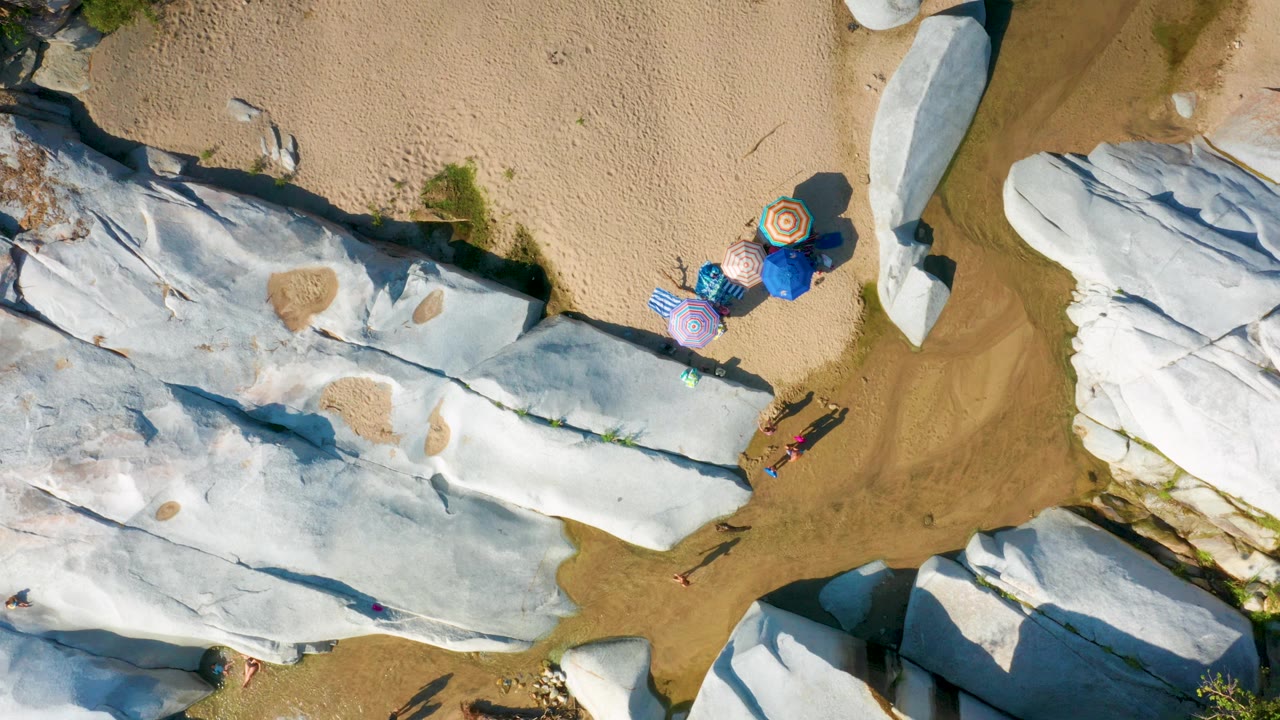 Aerial overhead frame in La Palma, Baja California Sur showing elongated pale rocks, tan beach, clustered umbrellas, and tiny visitors with long shadows near the rock margin.