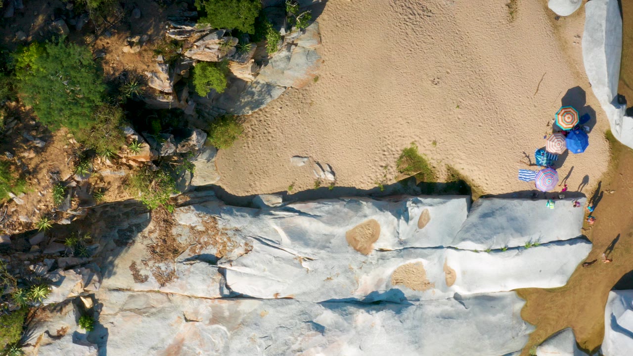 Top-down frame from La Palma, Baja California Sur showing pale white rock across tan sand, green shrubs at the left edge, and beach umbrellas near the upper-right side.