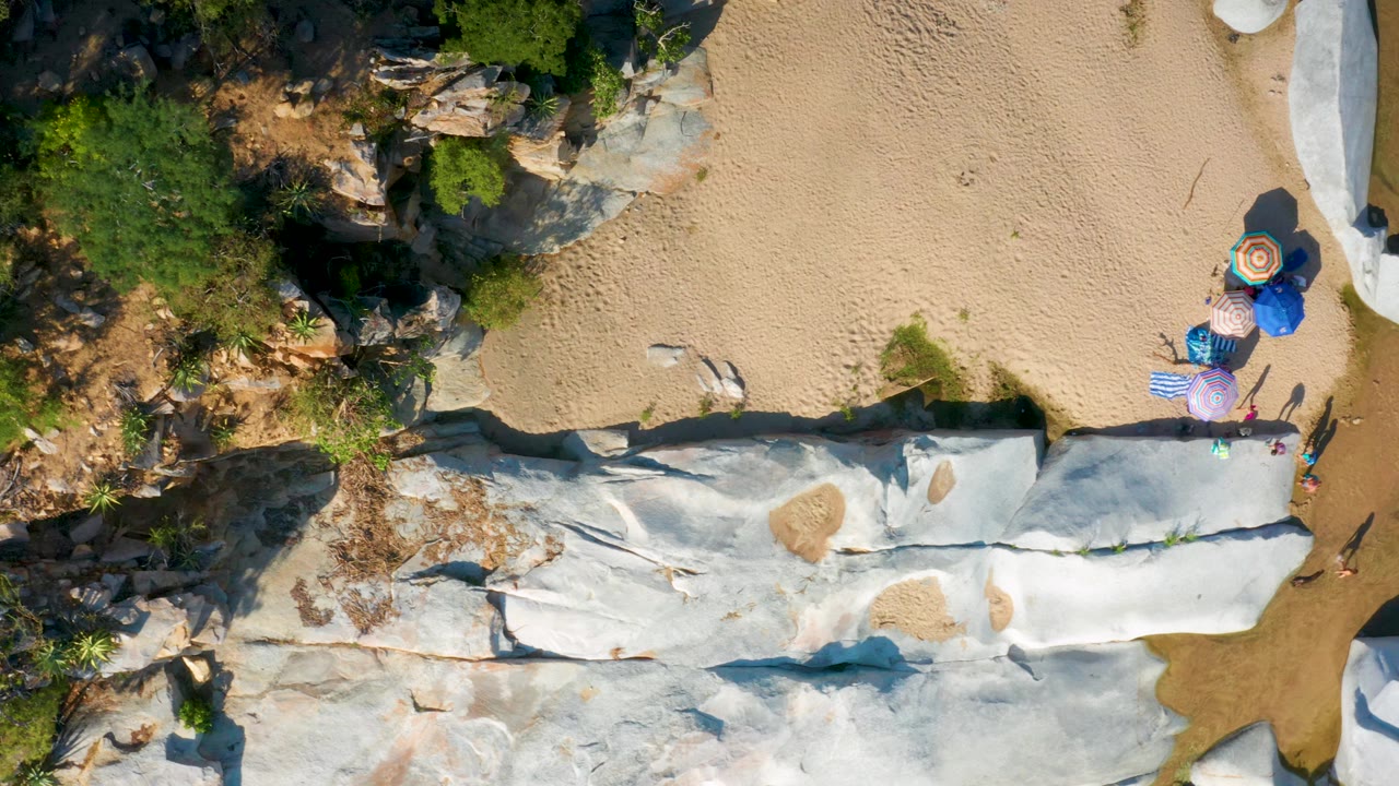A later overhead view in La Palma, Baja California Sur holds the pale rock ledge through the lower frame while tan sand and umbrellas remain near the upper-right edge.