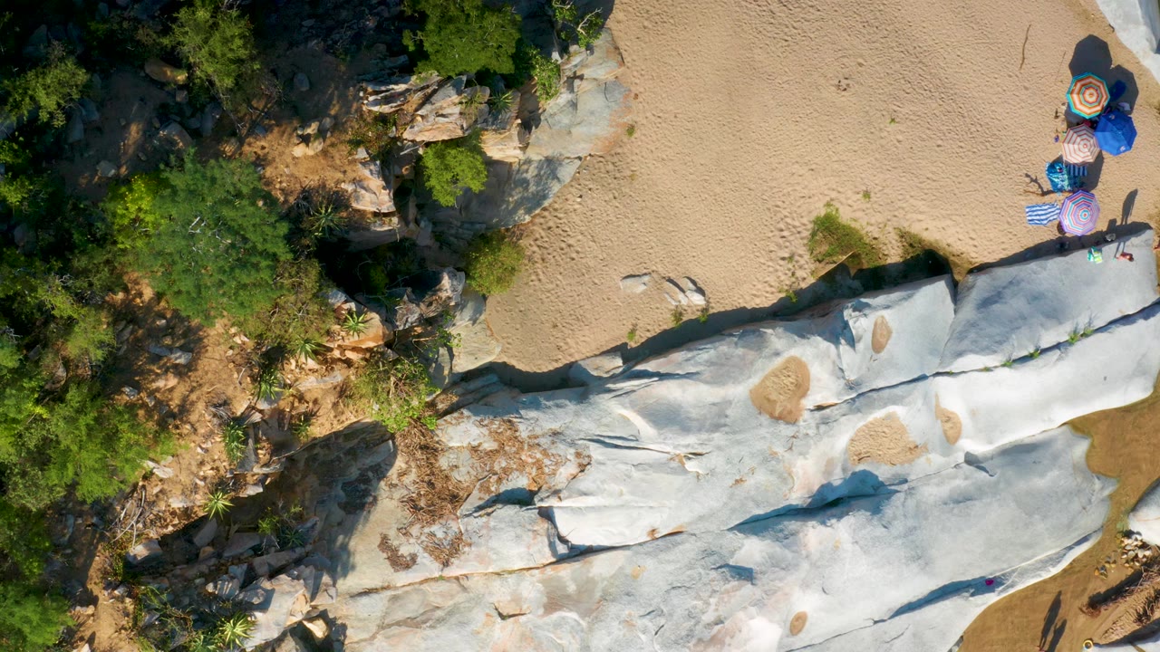 Mid-clip overhead frame from La Palma, Baja California Sur showing green vegetation along the left, pale rock across the bottom, tan sand above, and umbrellas near the right side.