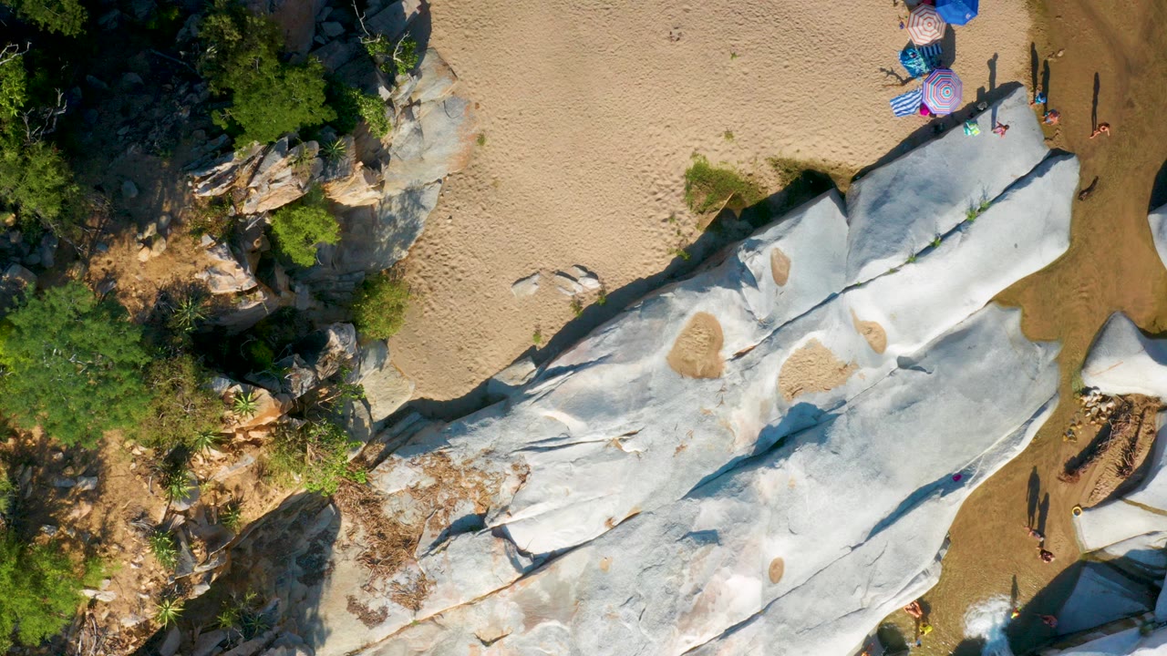 Overhead drone frame in La Palma, Baja California Sur showing pale rock slabs crossing tan sand, green brush on the left, umbrellas near the top edge, and several small visible people.