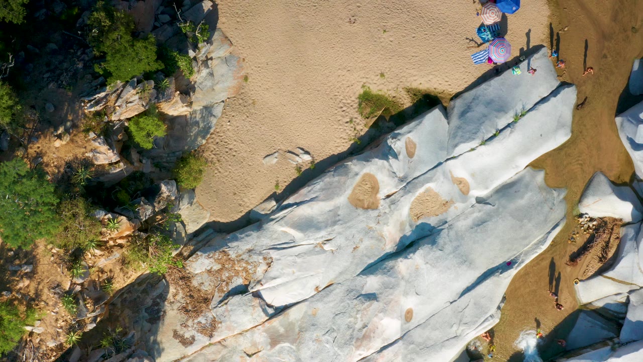 Aerial frame in La Palma, Baja California Sur showing pale stone ridges running diagonally through sand, with umbrellas and several visible people clustered near the upper right.