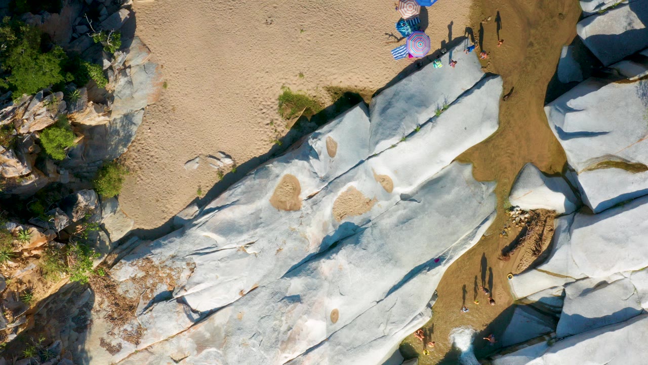 Overhead frame in La Palma, Baja California Sur showing pale rock slabs, tan beach sand, shallow water, umbrellas, and tiny visible tourists near the upper edge.