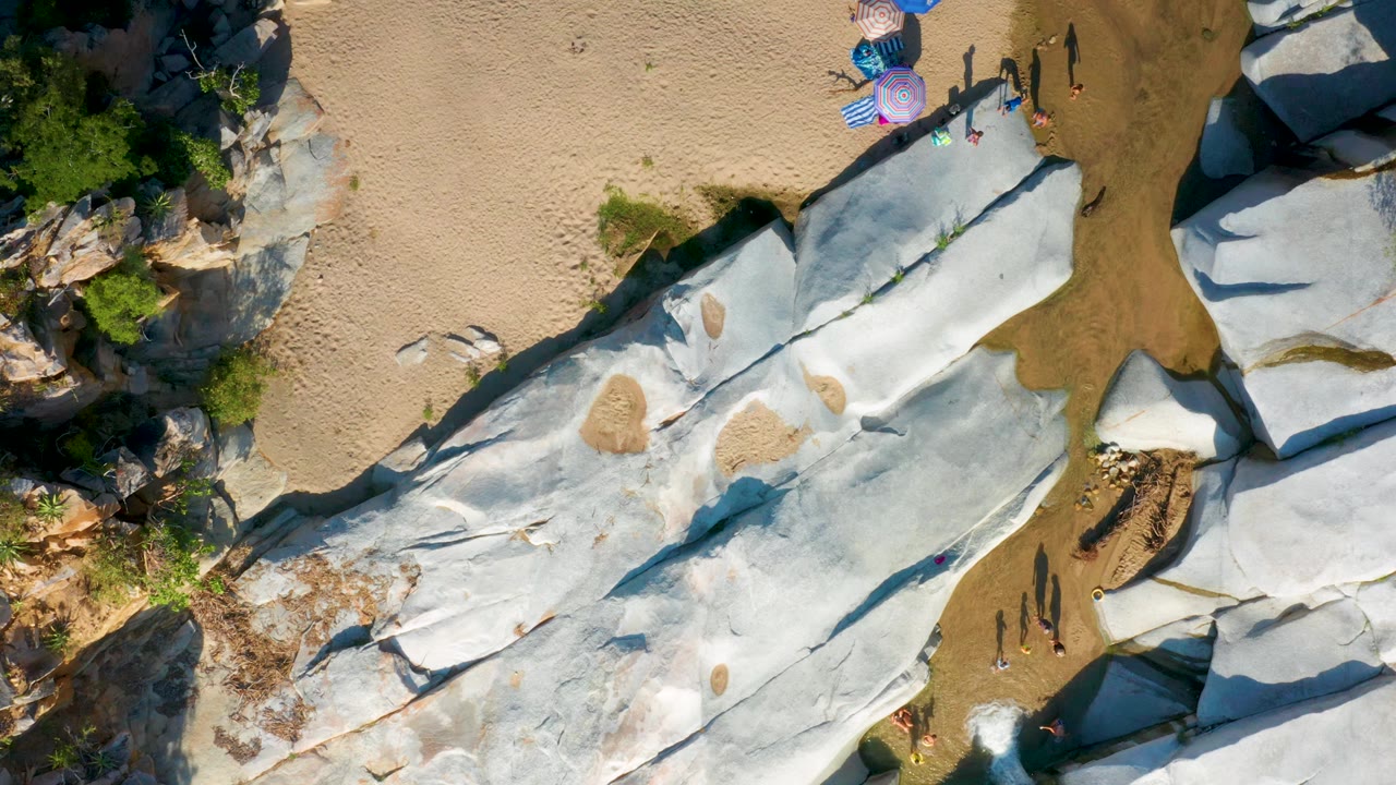 Aerial view of white-gray rock slabs with sand pockets, water, umbrellas, and tiny walking figures.