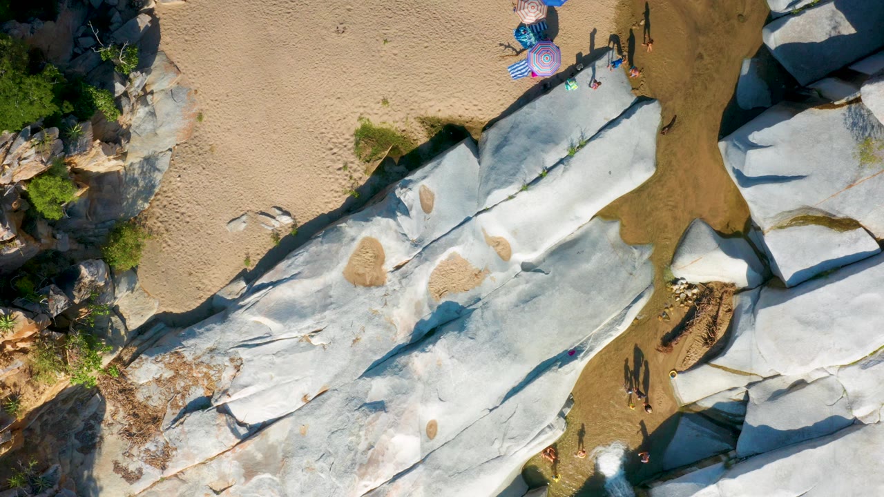 The aerial view in La Palma shows the pale stone formation stretching diagonally across frame, with tan sand, shallow brown-green water, umbrellas, and visible tiny tourists.