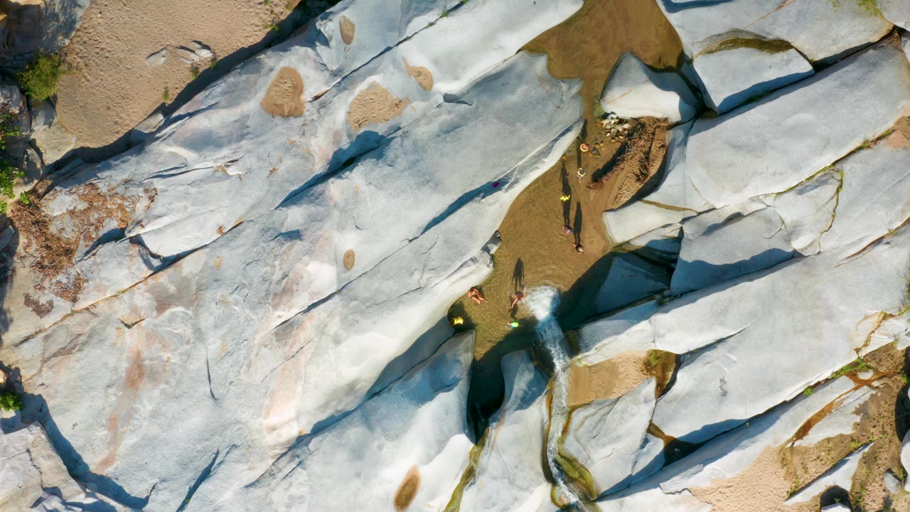 Overhead frame from La Palma, Baja California Sur showing pale rock slabs, tan sand pockets, a narrow water channel, and small people near the lower center.