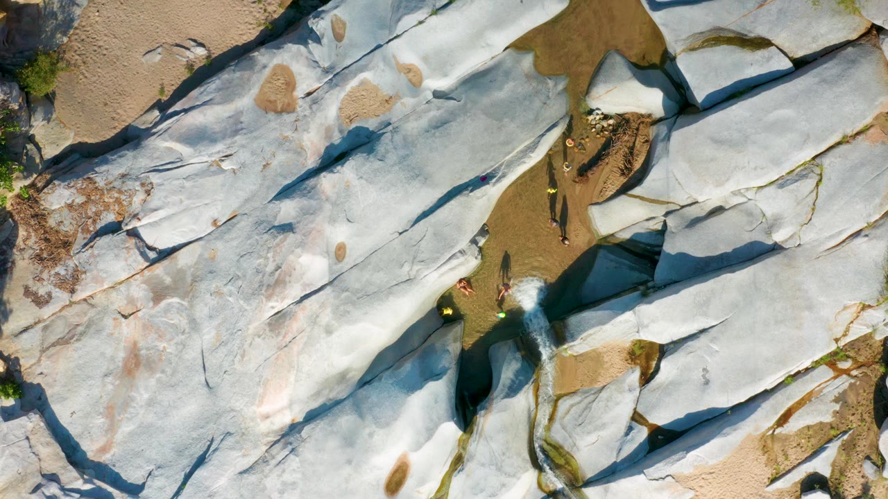 Aerial view in La Palma, Baja California Sur with white-gray rock plates crossing the frame, sand above and between them, and tiny people beside the water channel.