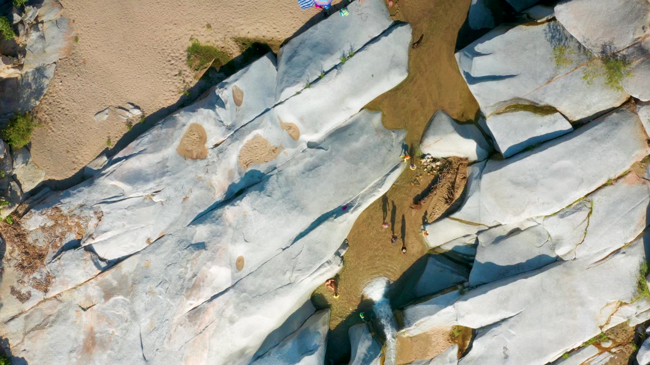 Top-down aerial frame from La Palma, Baja California Sur, showing pale rock shelves, tan sand, shallow brown water, sparse shrubs, and no clearly visible people.