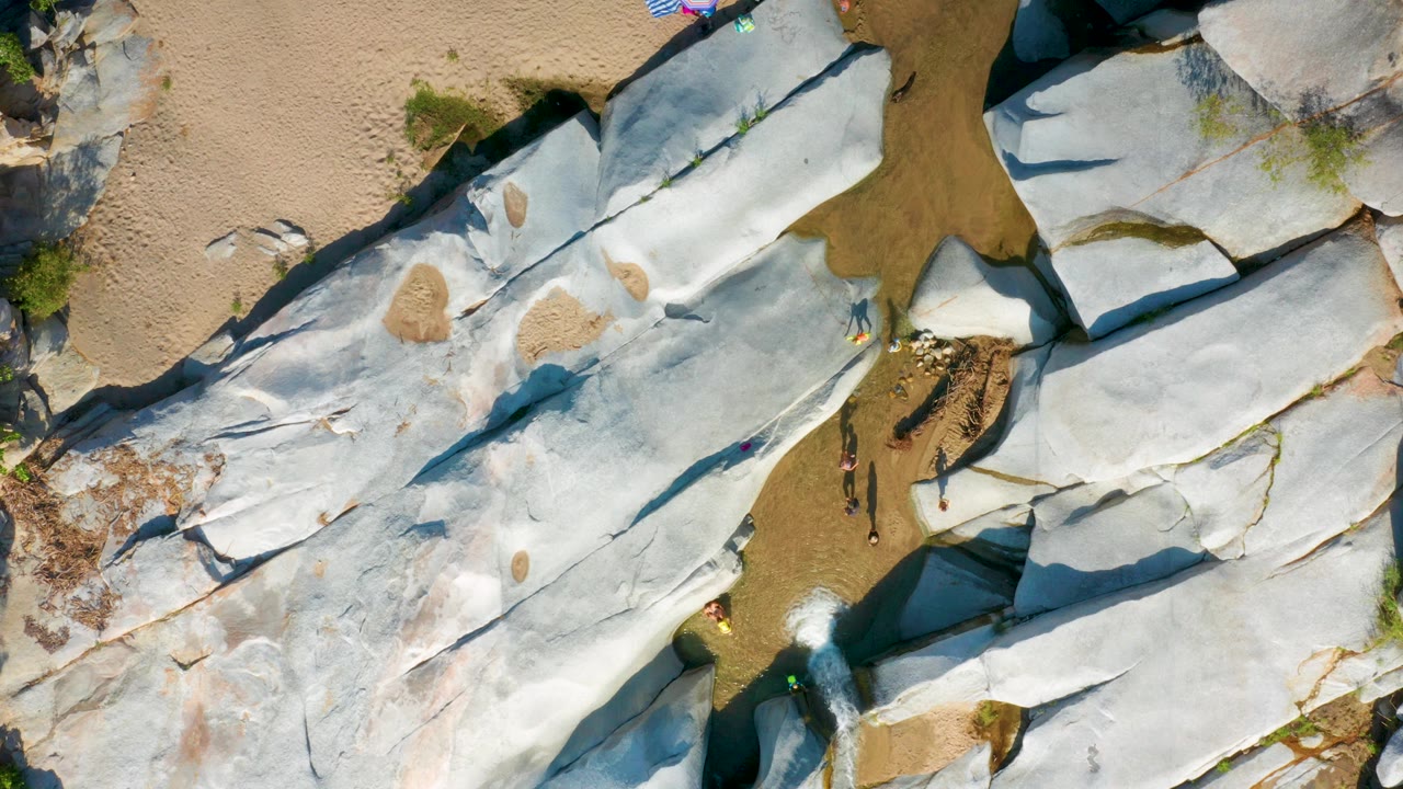 Overhead frame from La Palma, Baja California Sur, showing pale rock ledges around a shallow brown channel, tan sandy areas, small green plants, and no clearly visible people.