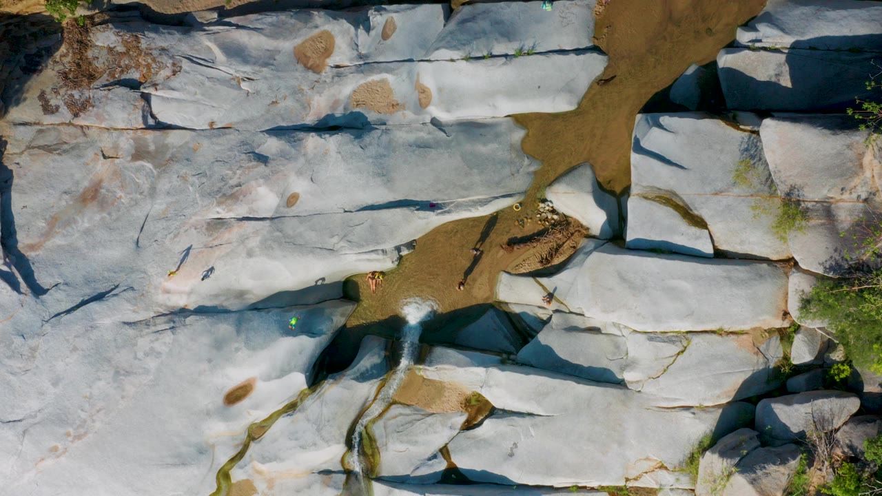 Top-down frame from La Palma, Baja California Sur showing pale white rock shelves crossed by brown sandy channels, edge shrubs, and tiny visible people near the wash.