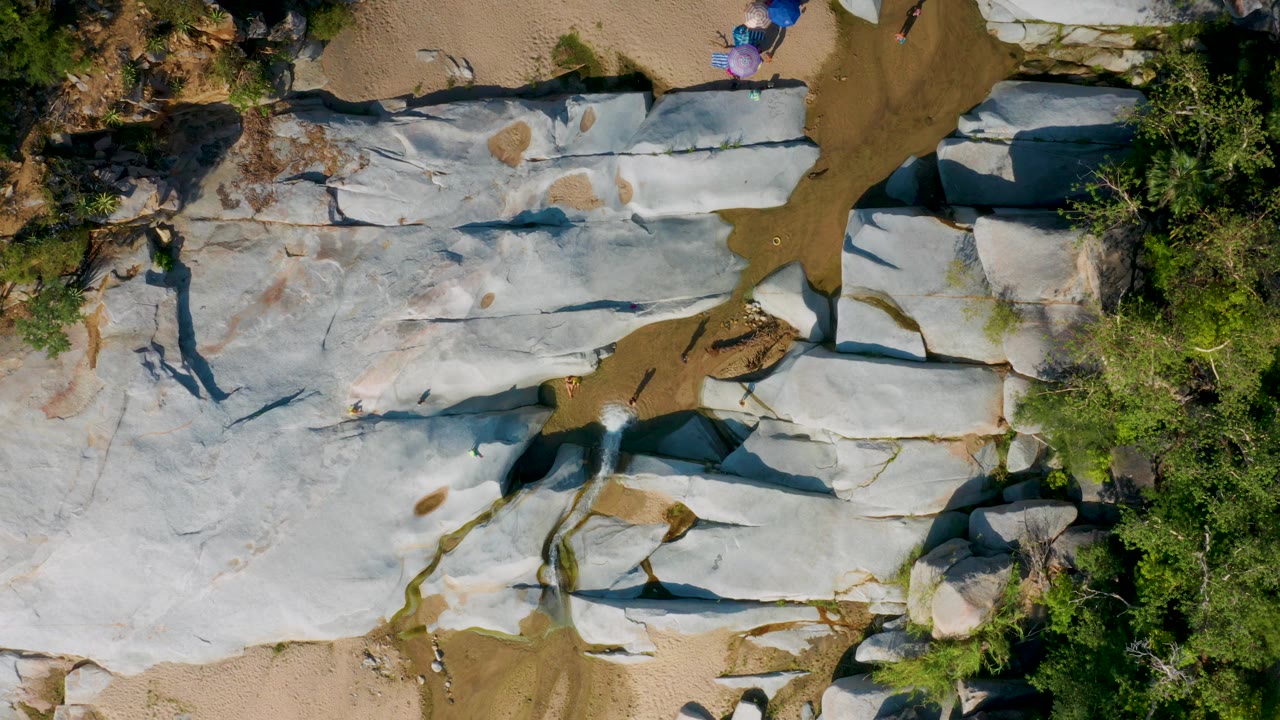 Top-down frame from La Palma, Baja California Sur with broad gray rock sheets crossing the center, sandy ground above, dense green trees at right, and small people near the top edge.