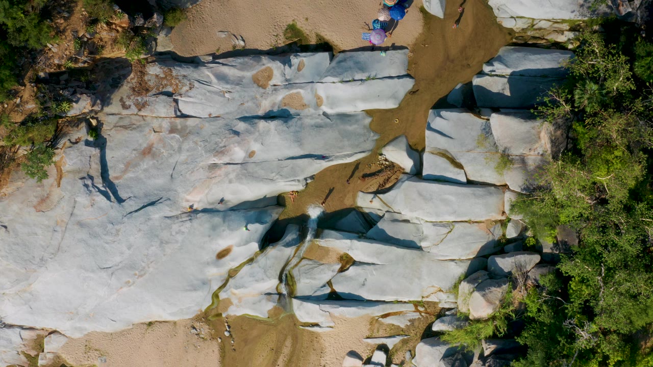 Aerial frame from La Palma, Baja California Sur showing white-gray rock forms cut by sandy channels, with green tree cover on the right and tiny people near the top border.