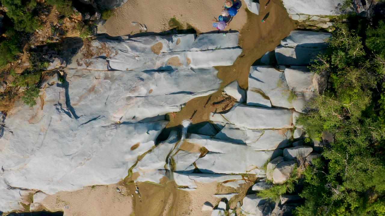 Overhead landscape of flat pale rock, tan sand, and trees with people near the upper right.