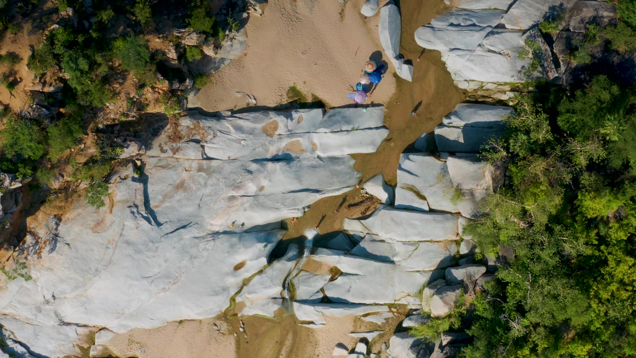 Overhead frame from La Palma, Baja California Sur showing pale rock slabs, tan sandy channels, dense green scrub at the right edge, and small people near blue and purple items.
