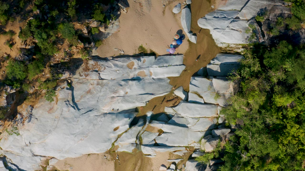 Aerial La Palma frame showing white and gray rock shelves on both sides of a tan sand channel, with thick green scrub in the upper frame and tiny people near the right-center rocks.
